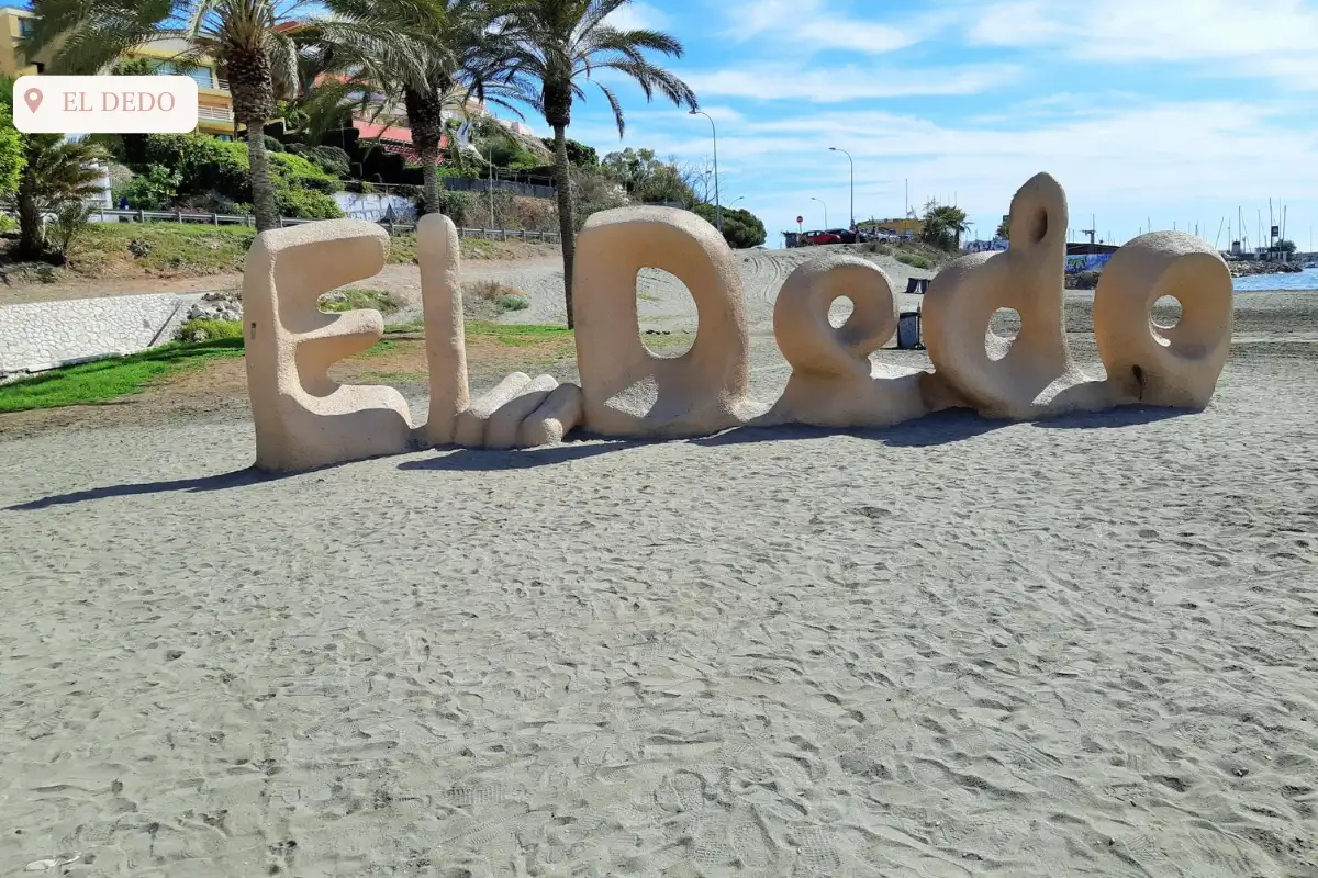 The El Dedo sign at Playa El Dedo on top of brown sand with a small port behind it.